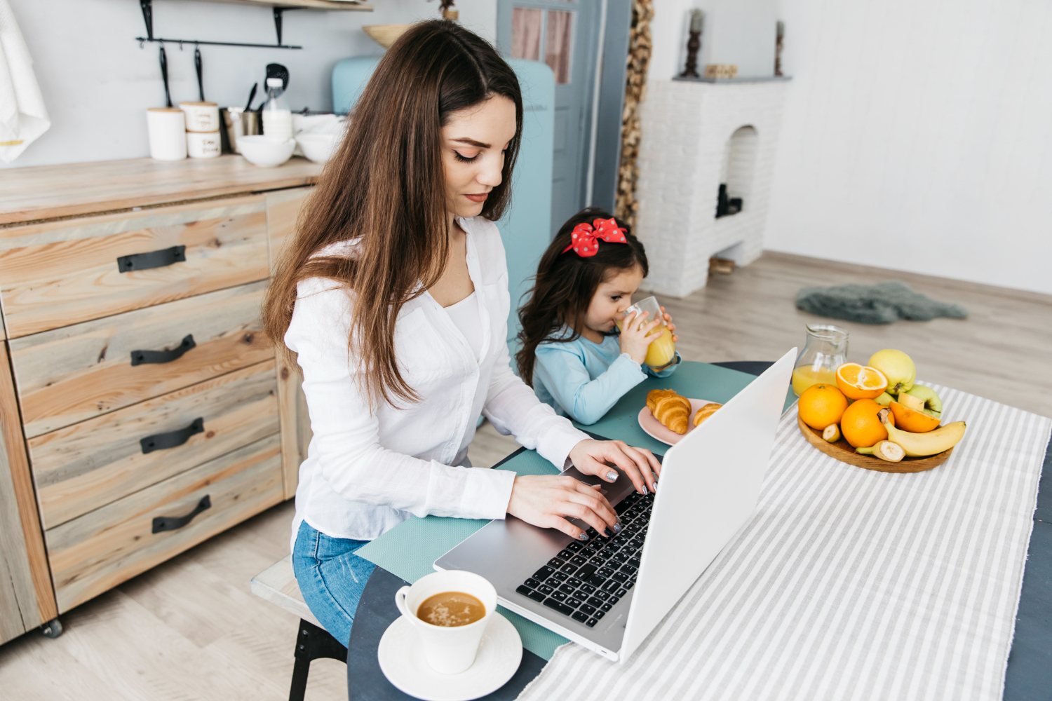Woman working on a laptop at the kitchen table while her young daughter eats breakfast beside her.