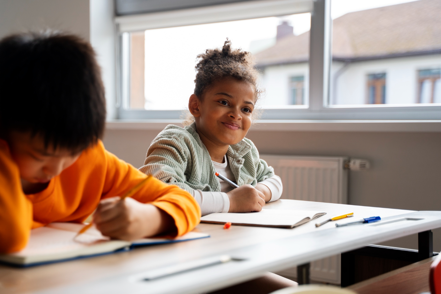 Two children sitting at desks in a classroom, writing in notebooks during a lesson.