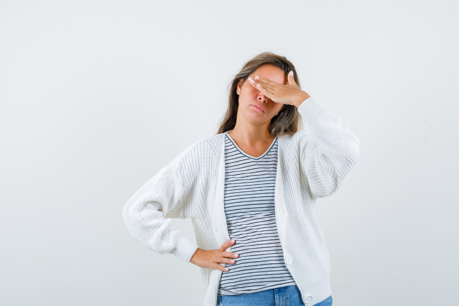 Tired woman covering her eyes with one hand while standing against a plain background.