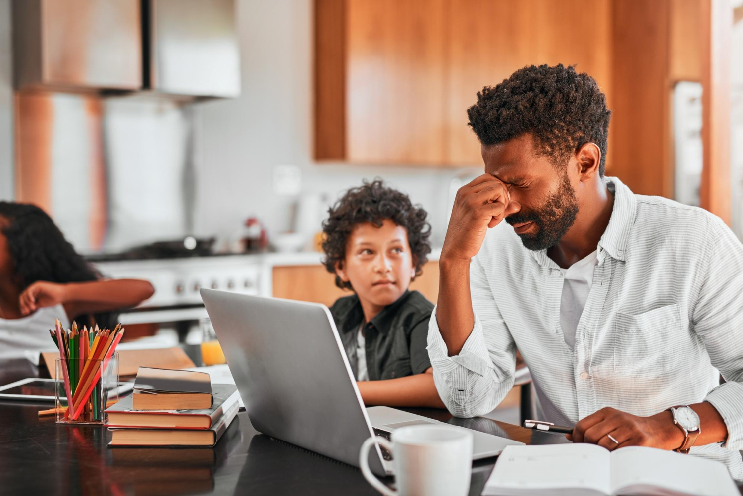 Stressed father working on a laptop at a kitchen table while children study nearby at home.