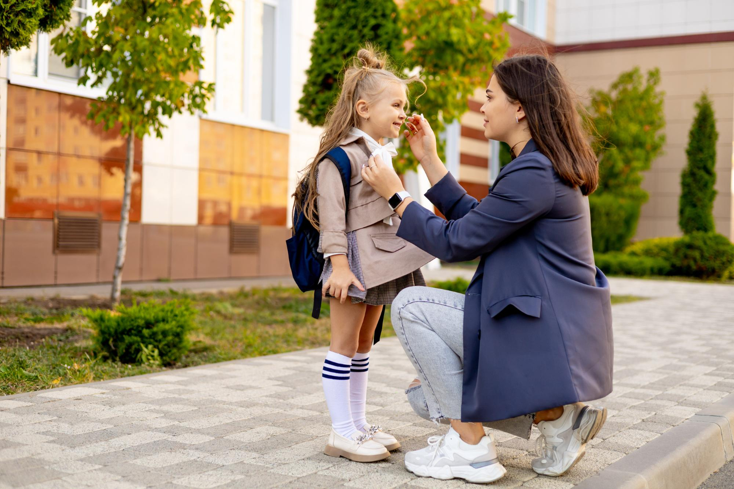 Mother kneeling outside a school, helping her young daughter adjust her outfit before class.