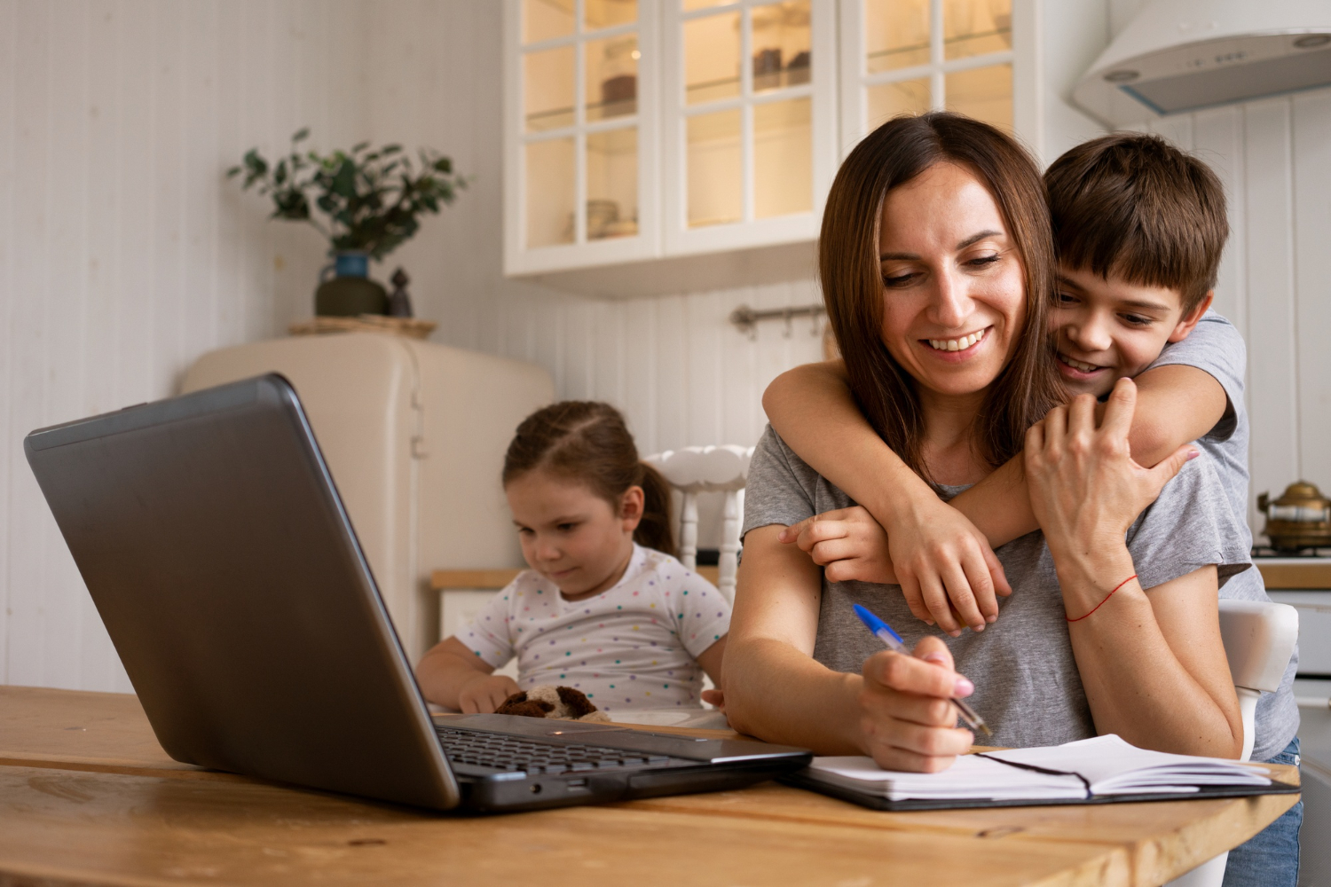Mother working at a laptop and writing in a notebook while her son hugs her and another child sits nearby at the kitchen table.