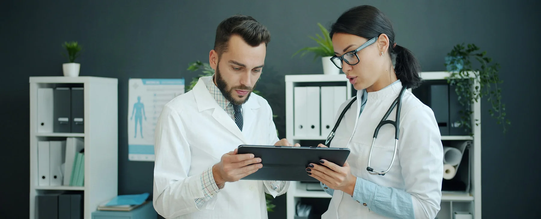 Two healthcare professionals in white lab coats stand in a medical office, reviewing information on a tablet together. One person wears a stethoscope and glasses, while both focus on the screen. Shelves with binders, plants, and medical materials are visible in the background.