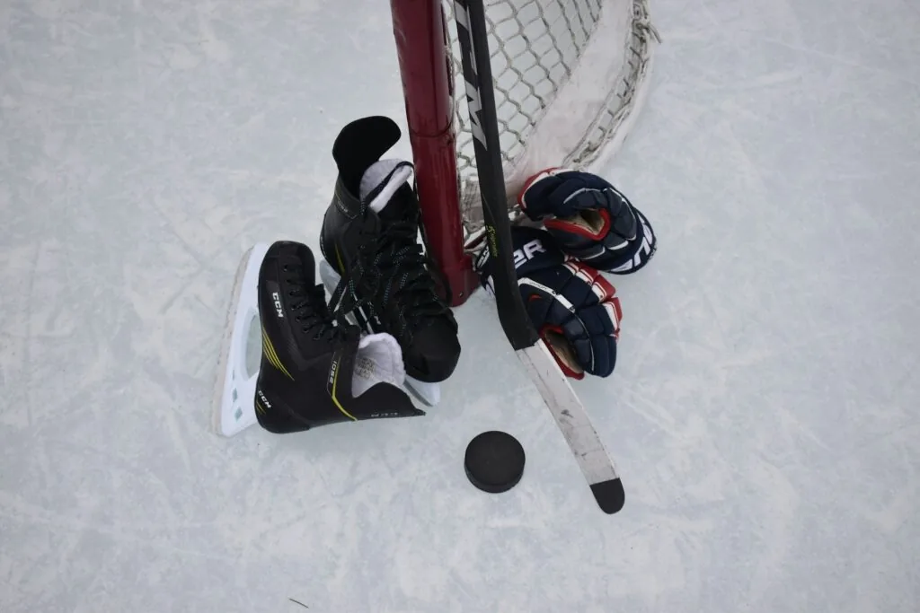 A pair of ice skates, hockey gloves, a hockey stick, and a puck are arranged on an outdoor ice rink against the post of a hockey goal.