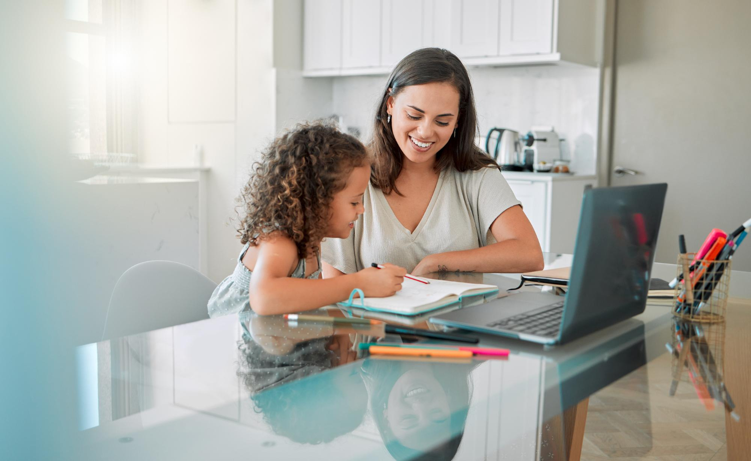 Mother and young daughter sitting at a kitchen table, smiling as they work together in a notebook beside a laptop.