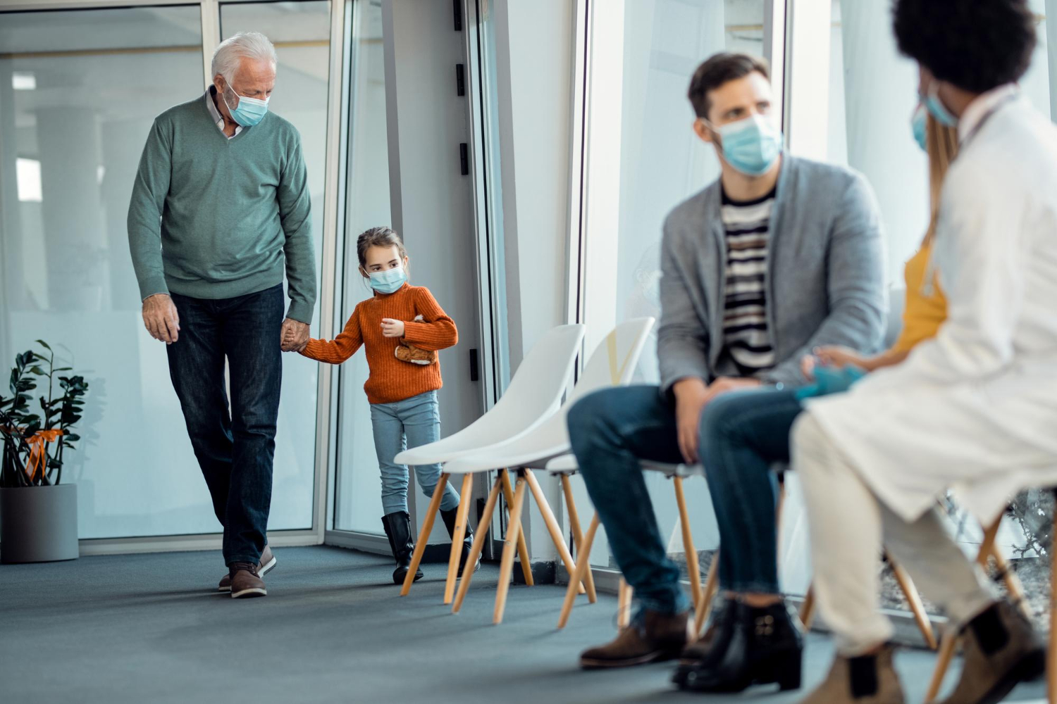 Older adult and child enter a healthcare waiting room while other patients wait to be seen, illustrating different patient experiences across generations.